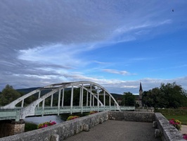 Eine neue Brücke über einen Fluss mit Blumenbeeten im Vordergrund und einem Kirchturm im Hintergrund.